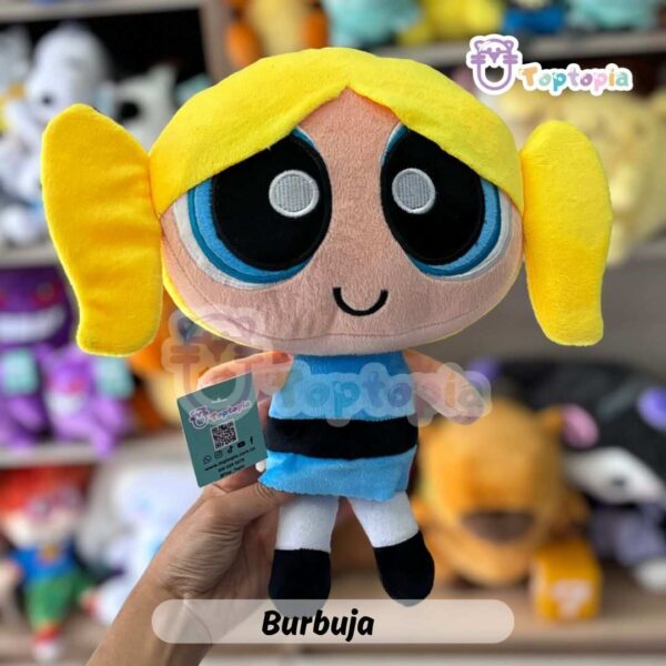 Peluche de Burbuja, personaje de Las Chicas Superpoderosas, con cabello amarillo, vestido azul y grandes ojos celestes, sobre fondo de tienda de juguetes.